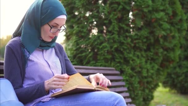 Μουσουλμανικές κορίτσι με τα γυαλιά, διαβάζοντας ένα βιβλίο σε ένα παγκάκι στο ηλιοβασίλεμα — Αρχείο Βίντεο