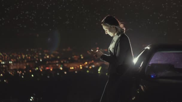 Retrato de una hermosa chica con un teléfono inteligente de pie junto al coche, luces de la ciudad por la noche — Vídeos de Stock