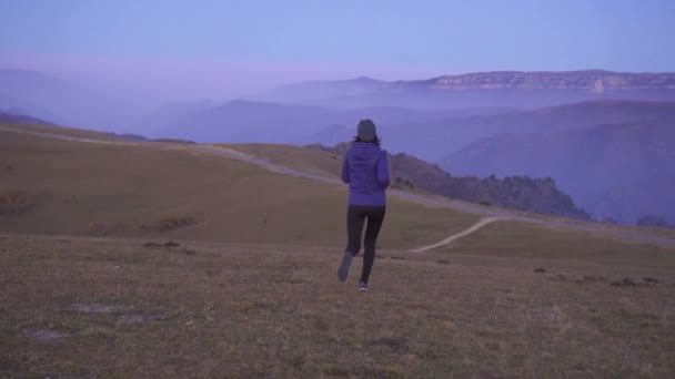Mladá žena sportovec v klobouku je spuštěn na pozadí hory, pohled zezadu, pomalé mo — Stock video