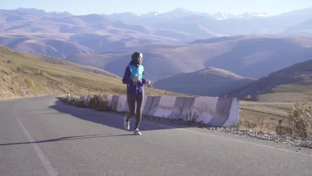 Giovane donna araba in un hijab corre lungo una strada di montagna soleggiata e si guarda intorno, lento mo — Video Stock