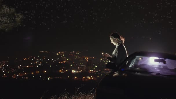 Retrato de una hermosa chica con un teléfono inteligente en el fondo de la ciudad de la noche y el cielo estrellado — Vídeos de Stock