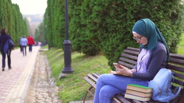 Estudiante musulmán con teléfono y libro en un banco del parque al atardecer — Vídeos de Stock