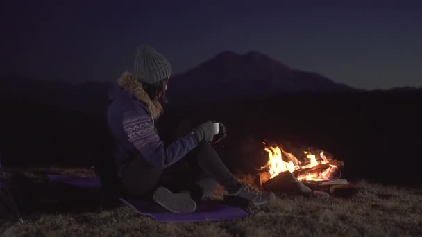 Kemping fiatal nő ül, és iszik egy pohár, a tűz körül éjjel a hegyekben, lassú mo — Stock videók