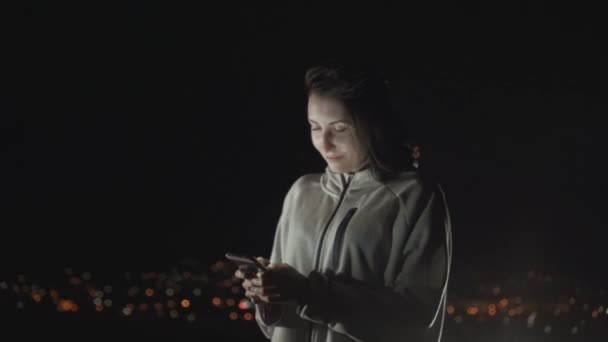 Ritratto di una ragazza con un telefono guarda nella fotocamera sullo sfondo della città di notte — Video Stock