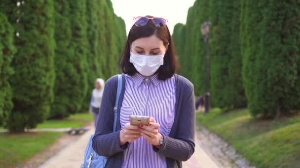Hezká dívka v ochranné lékařské maska na obličej v parku pomocí telefonu — Stock video