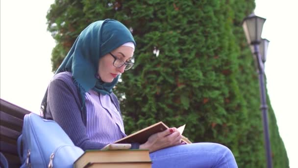 Мусульманская девушка в хиджабе читает книгу на коленях на скамейке в парке на закате — стоковое видео