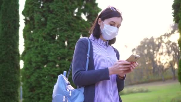 Meisje in een beschermende medische masker op haar gezicht gaat en maakt gebruik van de telefoon — Stockvideo