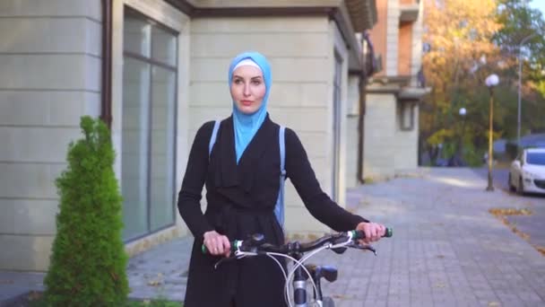 Мусульманка в хиджабе и с рюкзаком на велосипеде проходит через солнечный осенний город — стоковое видео