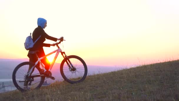 Mujer musulmana en un hiyab con una mochila con una bicicleta va cuesta arriba y mira la puesta de sol, lento mo — Vídeo de stock