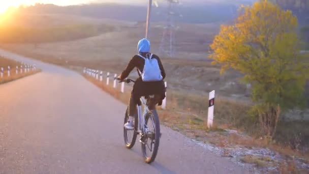 穆斯林妇女在盖头与背包骑自行车快速在道路上美丽的日落, 后视图 — 图库视频影像