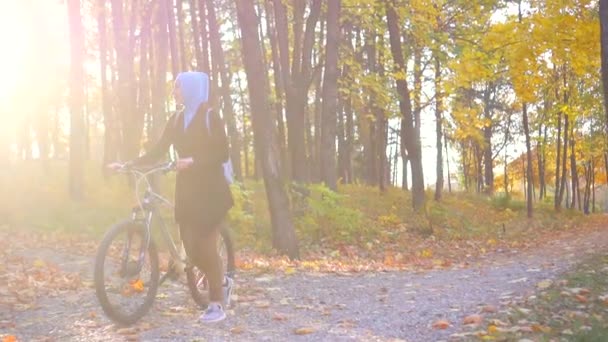 Mulher muçulmana em um hijab e com uma mochila com uma bicicleta passa por um parque de outono ensolarado, brilho do sol, câmera lenta — Vídeo de Stock