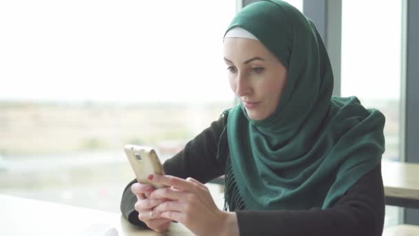 Porträt hübsche muslimische Frau im Hijab, die am Tisch sitzt und das Telefon benutzt, langsam — Stockvideo