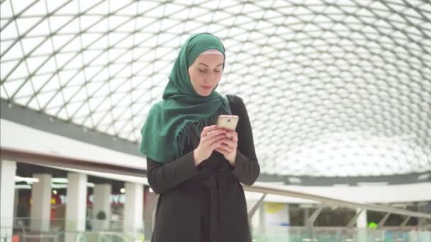 Mujer bastante musulmana en un hijab se para en un centro comercial y usa el teléfono — Vídeos de Stock