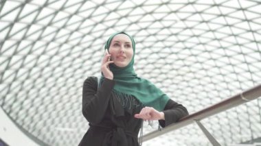 Bir sırt çantası ile bir hijab oldukça Müslüman kadında bir alışveriş merkezinde duruyor ve telefonda konuşurken, yavaş mo
