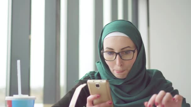 Jovem mulher muçulmana em um hijab está sentado a uma mesa pela janela usando o telefone e comer batatas — Vídeo de Stock