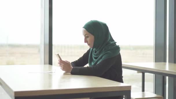Красивая мусульманка в хиджабе сидит за столом и пользуется телефоном, медленное время — стоковое видео