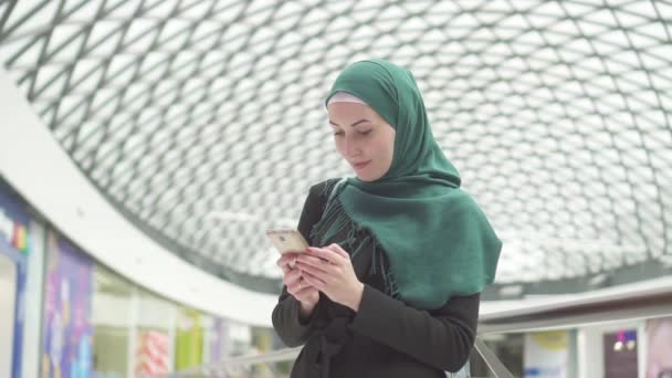 若いイスラム教徒の女性 hijab を厳しく身にショッピング センターに立っているし、遅い mo、携帯電話を使用して — ストック動画