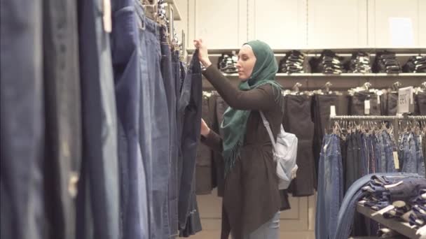 Mulher muçulmana no hijab e uma mochila escolhe jeans na loja no shopping — Vídeo de Stock