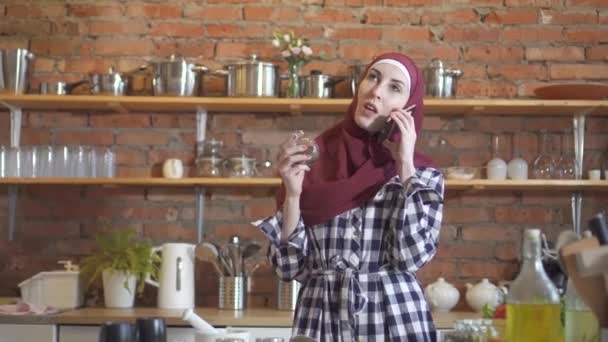 Νεαρή μουσουλμάνα μιλώντας στο τηλέφωνο και την προετοιμασία φαγητού στην κουζίνα — Αρχείο Βίντεο