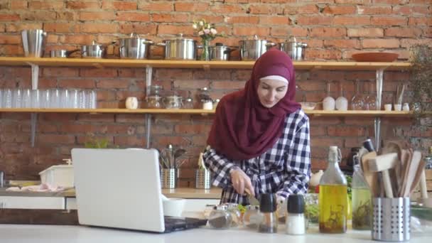 Μουσουλμανική γυναίκα σε ένα hijab στην κουζίνα βλέπουν ένα βίντεο κατάρτισης φορητό υπολογιστή πώς να μαγειρεύουν ένα πιάτο — Αρχείο Βίντεο