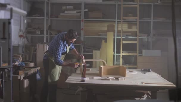 Cabinet maker in a shirt with a beard in a dark autotechnoy workshop paints a tree — Stock Video