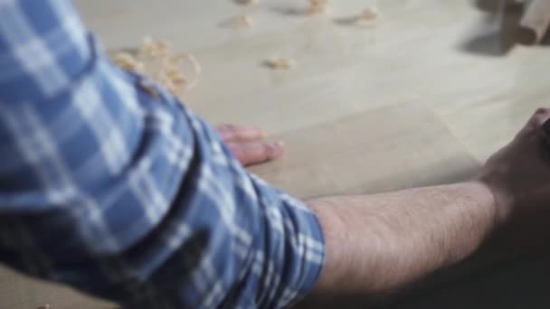 Τα χέρια ενός ξυλουργού σε ένα πουκάμισο σε ένα εργαστήριο με ένα αεροπλάνο πετάει ροκανίδια, κοντινό πλάνο — Αρχείο Βίντεο
