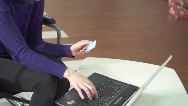Giovane donna disabile con gli occhiali sul sedile della sedia a rotelle con un computer portatile a casa, utilizza una carta bancaria — Video Stock