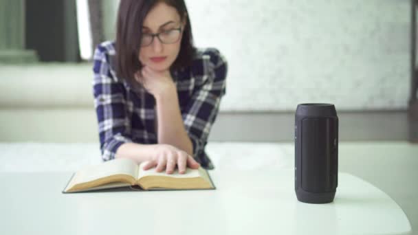 Hermosa chica con gafas utiliza asistente de voz al leer un libro — Vídeos de Stock