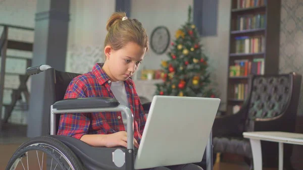 Teenage girl disabled in a wheelchair, sitting at the laptop, against the Christmas tree Royalty Free Stock Photos