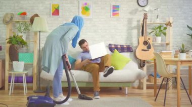 Bir başörtüsü içinde Müslüman kadın modern bir daire, kanepede oturan ve bir akıllı telefon kullanarak genç bir adam vakumlama