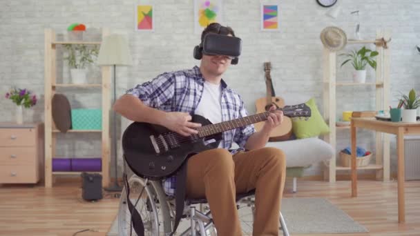 Portret młody człowiek niepełnosprawny w pewien wózek inwalidzki w czynny rzeczywistość okulary muzyk gra ten elektryczny gitara — Wideo stockowe