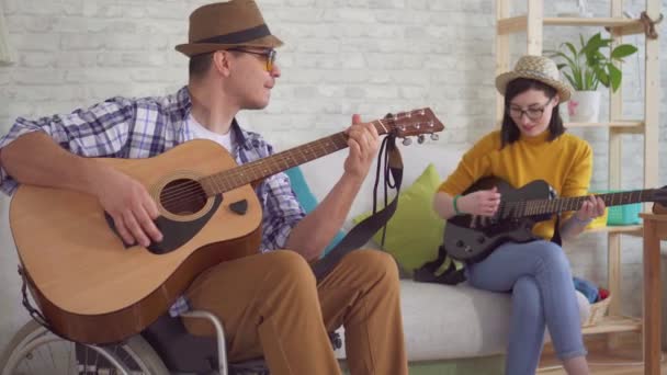 Молодой человек - инвалид-музыкант в инвалидной коляске, девушка в очках и шляпа, играющая на гитаре. — стоковое видео