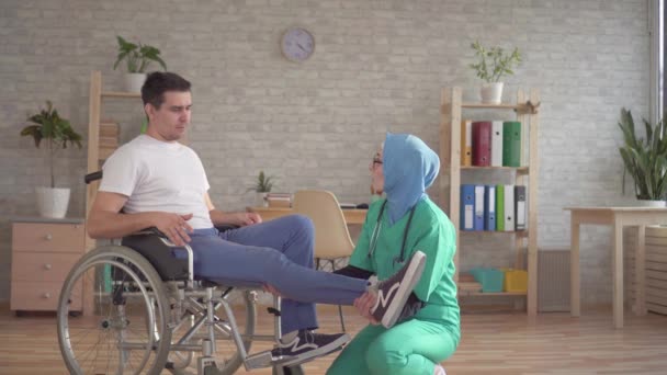 Moslim arts in hijab onderzoekt het been van een gehandicapte persoon in een rolstoel — Stockvideo