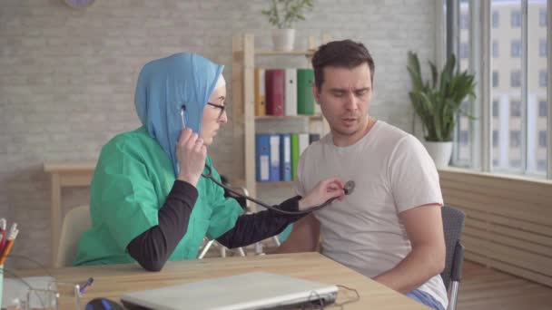 Médico muçulmano em hijab usando um estetoscópio ouve o batimento cardíaco de um paciente do sexo masculino — Vídeo de Stock