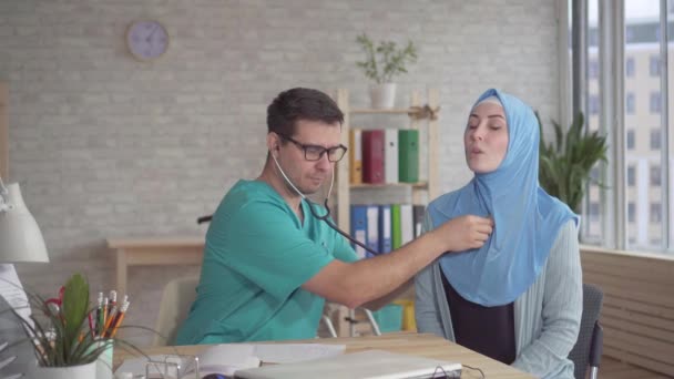 Bir stetoskop kullanan genç erkek doktor bir hijab genç bir Müslüman kadının kalp atışı dinler — Stok video