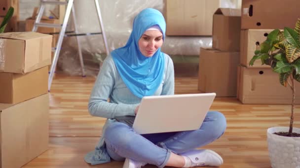 Retrato jovem muçulmano mulher em um hijab usa um laptop sentado no chão ao lado das caixas — Vídeo de Stock