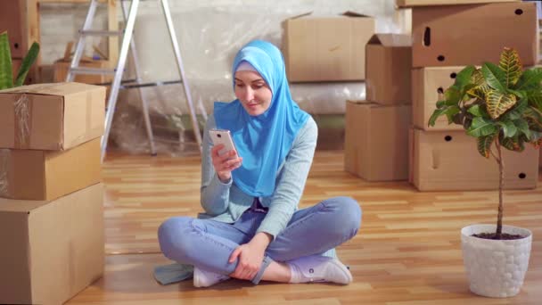 Portret jonge moslimvrouw in een hijab maakt gebruik van smartphone zittend op de vloer naast de dozen in een modern appartement — Stockvideo