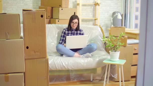 Jovem mulher alegre usa laptop sentado no sofá depois de se mudar para um apartamento moderno — Vídeo de Stock