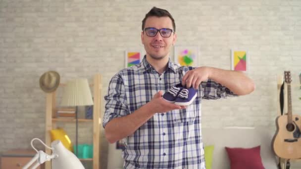 Νέος άντρας με γυαλιά κρατώντας παιδικά παπούτσια και κοιτάζοντας την κάμερα — Αρχείο Βίντεο