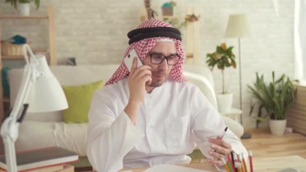 Homem árabe sentado à mesa e falando em um smartphone — Vídeo de Stock