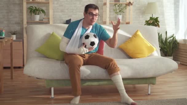 Expressive young man with a broken arm and leg sitting on the couch with a soccer ball learns about win — Stock Video