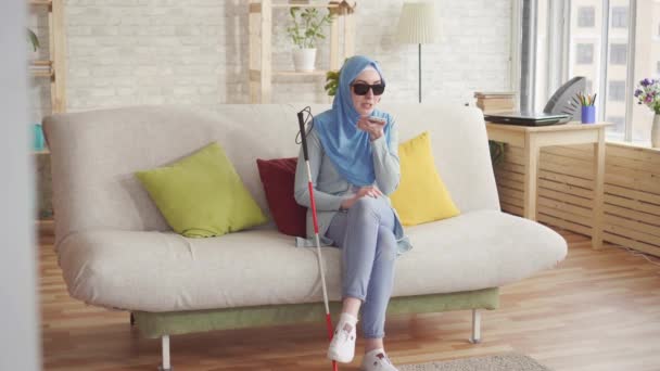 Mooi blind moslim meisje met een stok in hijab thuis, geniet van smartphone zittend op de Bank — Stockvideo