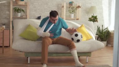 Kanepede oturan kırık bir kol ve bacak ile Expressive genç adam kazanmak yavaş mo hakkında öğrenir