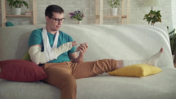 Porträt eines jungen Mannes mit gebrochenem Arm und Bein, der auf einem Sofa sitzt und ein Smartphone benutzt — Stockvideo