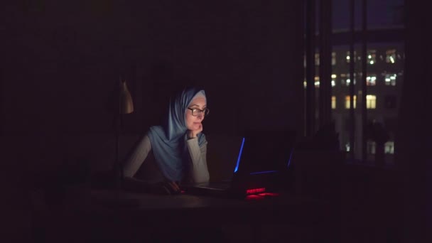 Ragazza musulmana con gli occhiali e un hijab con notebook nella ricerca scura su Internet a casa — Video Stock