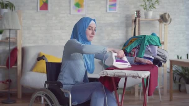 Gyönyörű muzulmán nő fogyatékkal élő kerekesszékkel, csinál házi feladatot, vasalás ruhák — Stock videók