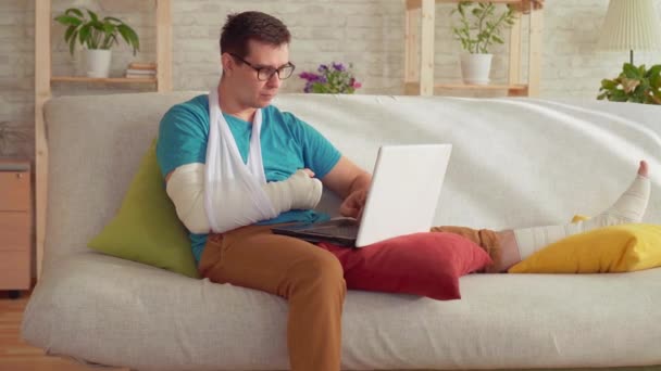 Retrato de un joven con un brazo y una pierna rotos sentado en el sofá y utiliza una computadora portátil — Vídeo de stock