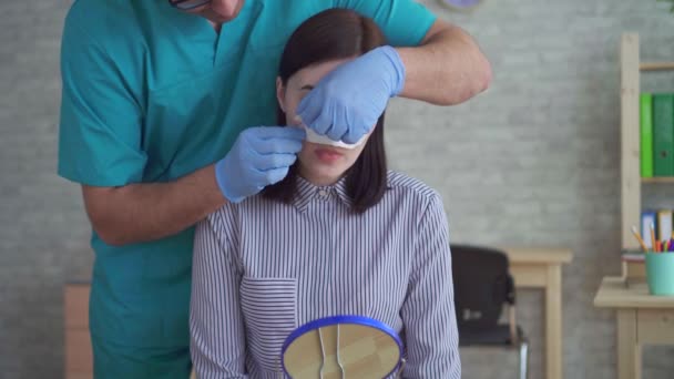남자 의사 성형 외과 의사는 수술 후 여성의 얼굴에서 붕대를 제거 — 비디오