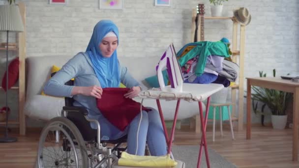 Όμορφη μουσουλμανική γυναίκα απενεργοποιημένη σε αναπηρικό καροτσάκι, κάνοντας εργασία, σιδέρωμα ρούχων — Αρχείο Βίντεο