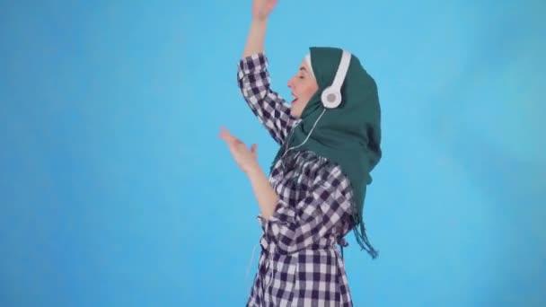 Porträt einer jungen muslimischen Frau mit Kopfhörern, die Musik hört und auf blauem Hintergrund tanzt — Stockvideo